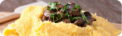 Professzionális kulináris fotó, közeli felvétel, „food-style” stílusban. A képen egy étel látható: sűrű, sárga polenta, amelyet halomként tálaltak egy fa vágódeszkára. A polentát tetején bőségesen díszítik pirított, barna gombák és apróra vágott friss zöld petrezselyem.